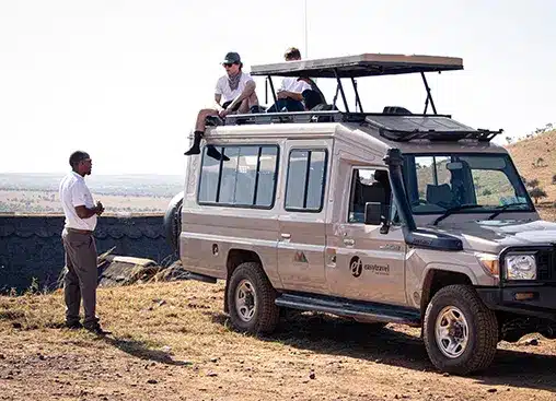 Een safarivoertuig met zitplaatsen op het dak op een savanne, twee personen bovenop, een gids die er vlakbij staat, een heldere hemel op de achtergrond.
