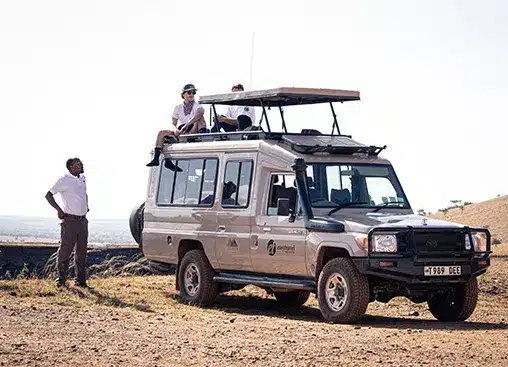Véhicule de safari avec sièges sur le toit dans la savane, trois personnes observant la faune sauvage, le guide se tenant à proximité.