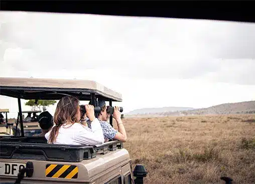 ジープに乗ったサファリ観光客が、曇り空の下の広いサバンナで双眼鏡を使って野生動物を観察しています。.