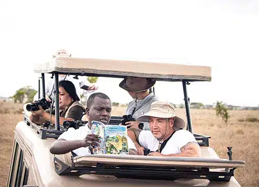 Safari tourists in an open-top vehicle observing wildlife, with one man holding a guidebook, in a savannah landscape.