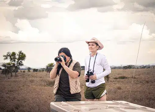 Due persone in un safari, una con una macchina fotografica e l'altra con un binocolo, in un paesaggio erboso di savana.
