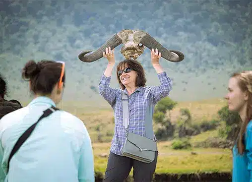 Une femme tenant un crâne d'éléphant dans un paysage herbeux, entourée de personnes, illustrant l'exploration de la faune sauvage.