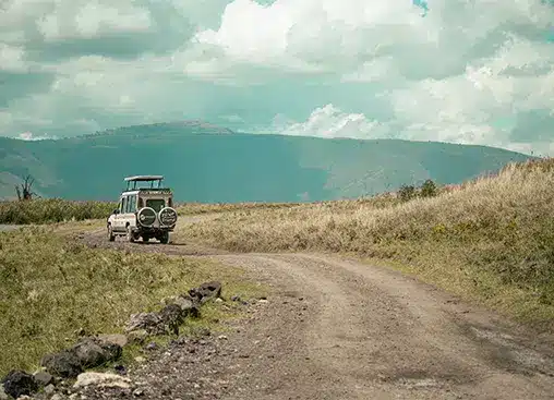 Внедорожник на грунтовой дороге посреди обширной травянистой местности с холмами вдалеке и облачным небом.