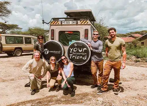 Grupp på sex personer poserar vid ett safarifordon i Tanzania, med reservdäcksskydd med texten "Din tanzaniska vän".