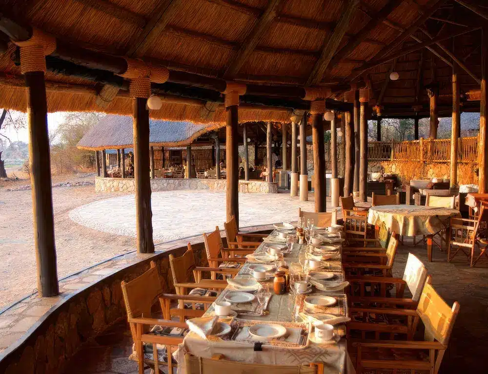 Eetgelegenheid in Ruaha River Lodge, Tanzania, met rustieke houten tafels, stoelen en een rieten dak met uitzicht op de rivier...