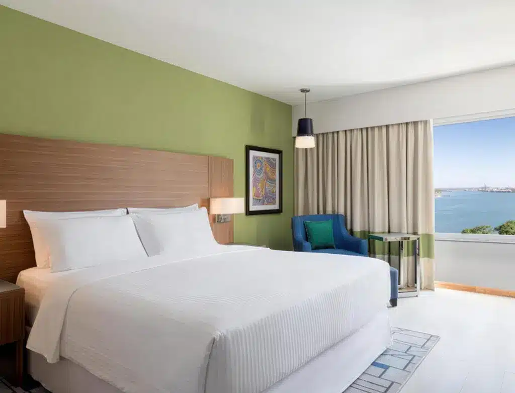 Modernes Hotelzimmer mit Kingsize-Bett, grüner Akzentwand, Kunstwerken, blauem Sessel und Meerblickfenster im Four Points...