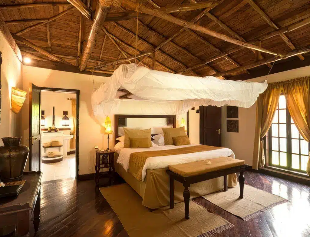 棕榈桑给巴尔酒店温馨的卧室，采用木质装饰，配有四柱床、暖色调灯光，敞开的房门通向明亮宜人的室外空间…….