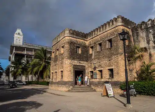 Historic stone building with tourists at entrance, palm trees, and a clock tower under a cloudy sky.