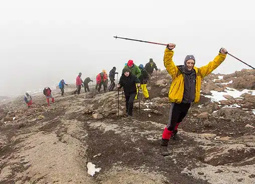 Grupo de excursionistas con chaquetas de colores caminando por un sendero rocoso en una montaña, con una persona celebrando en primer plano.