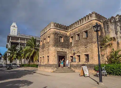 Historisch stenen gebouw met toeristen bij de ingang, palmbomen en een klokkentoren op de achtergrond onder een bewolkte hemel.