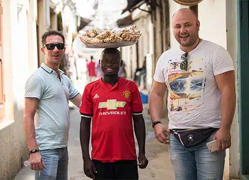 Drei Männer in einer engen Straße, einer trägt einen Korb auf dem Kopf, lächelnd und posierend für ein Foto.