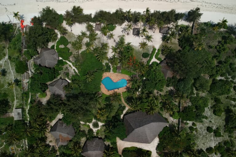 Luftaufnahme eines tropischen Resorts mit üppigem Grün, einem zentralen Pool und strohgedeckten Hütten in der Nähe eines Sandstrandes.