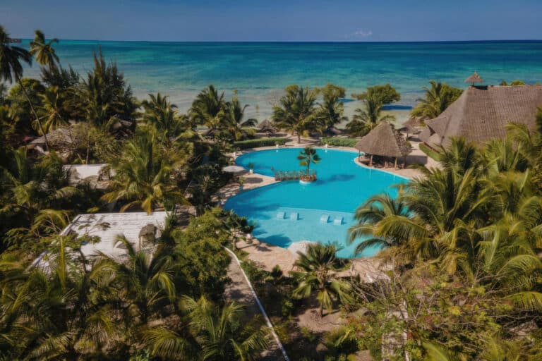 Resort tropical con palmeras, una gran piscina y vista al mar bajo un cielo azul claro.