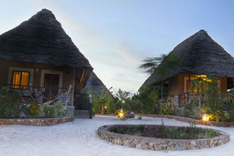 Strohgedeckte Bungalows am Sandstrand bei Sonnenuntergang, umgeben von tropischen Pflanzen und sanfter Beleuchtung.