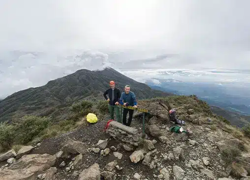 Two hikers stand at a mountain summit with a scenic view, cloudy sky, and rugged terrain, holding a sign.