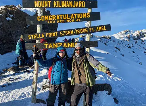 Twee wandelaars vieren feest bij Stella Point op de Kilimanjaro, omringd door sneeuw en een bord dat de hoogte van 5756 meter in Tanzania aangeeft.