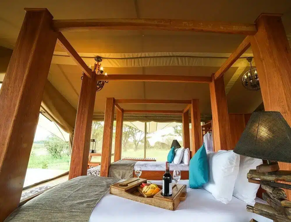 Luxuriöses Safari-Zelt-Interieur im Asanja Grumeti mit einem gemütlichen Bett, Holzelementen und einem Tablett mit Wein und Snacks.