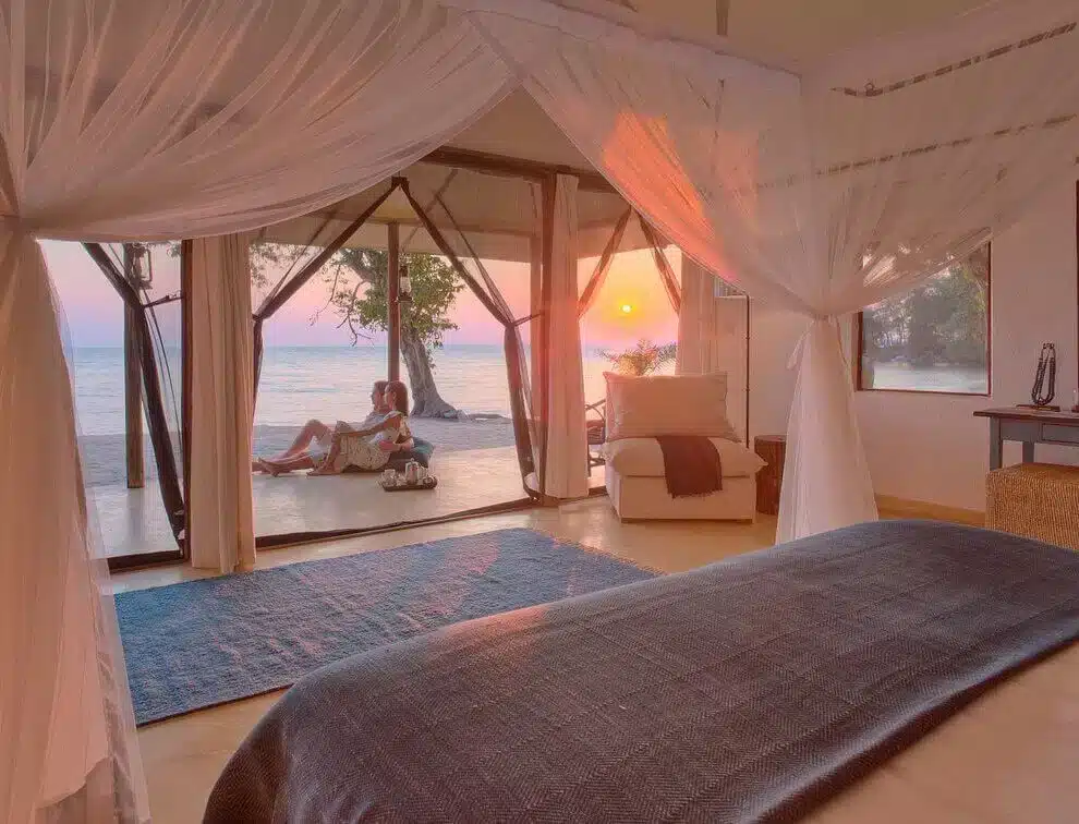 Lussuosa camera al Rubondo Island Camp con letto a baldacchino e vista su un sereno tramonto sul lago attraverso le porte a vetri aperte.
