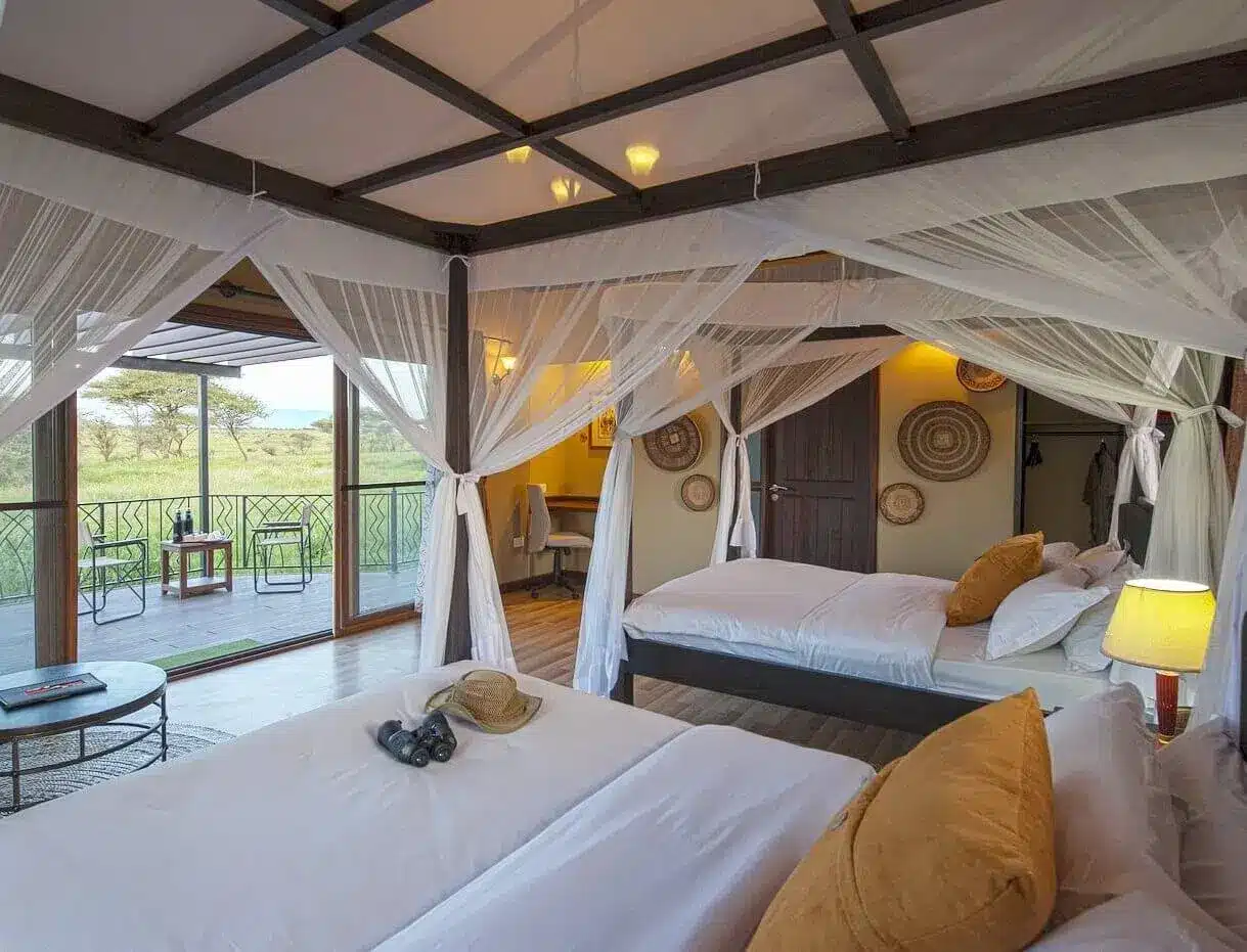 Chambre luxueuse dans un camp de safari, avec lits à baldaquin, moustiquaires et vue sur le paysage du Serengeti à travers des portes vitrées.