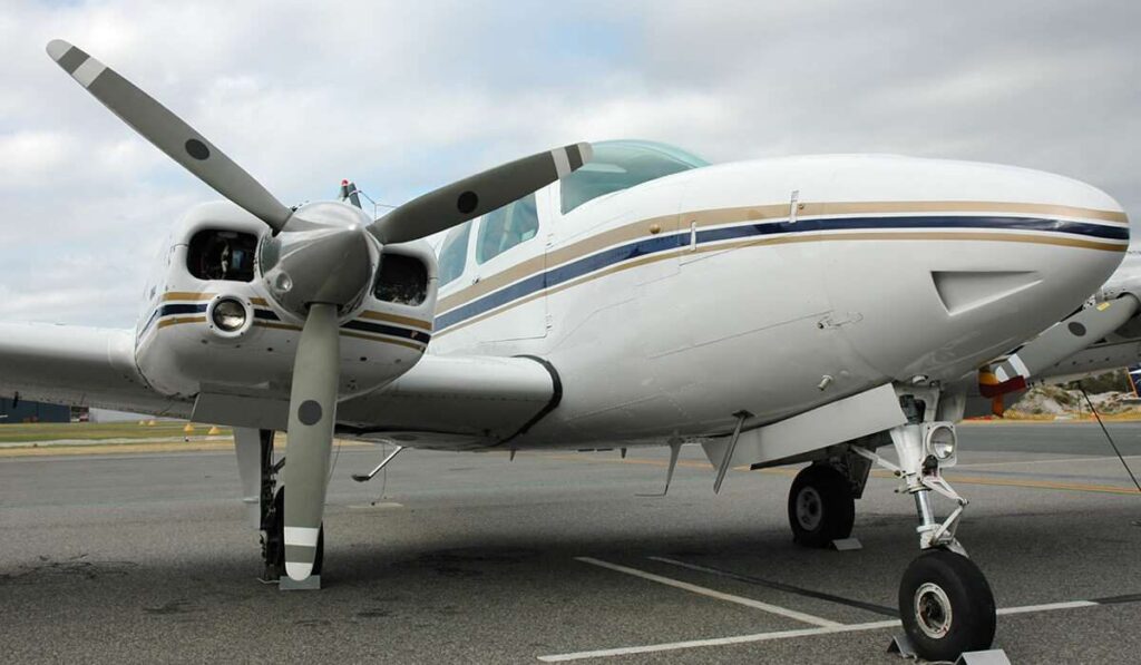 青と金のストライプが入った白い小型プロペラ機がアスファルト路面に駐機している。機体の前部とプロペラが見え、背景には曇り空が広がっている。 - easy travel tanzania