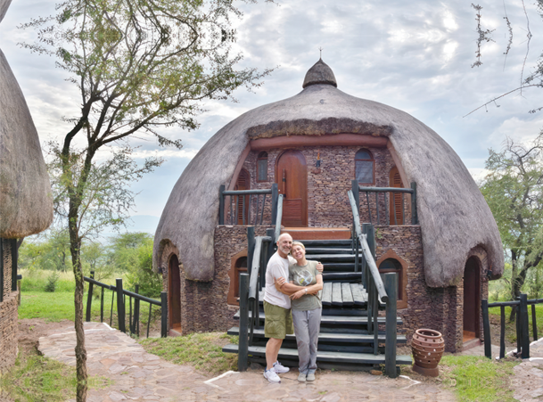 Een stel omhelst elkaar buiten een traditionele rieten hut bij Serengeti Serena Lodge in Tanzania, omgeven door weelderig groen en de hemel.