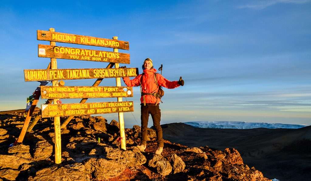 Eine Person posiert triumphierend neben einem Gipfelschild nach dem Aufstieg auf den Kilimandscharo. Das Schild zeigt Höhen und Orte an, mit felsigem Gelände im Vordergrund und einem Panoramablick auf die Berge unter einem klaren blauen Himmel im Hintergrund. - easy travel tanzania