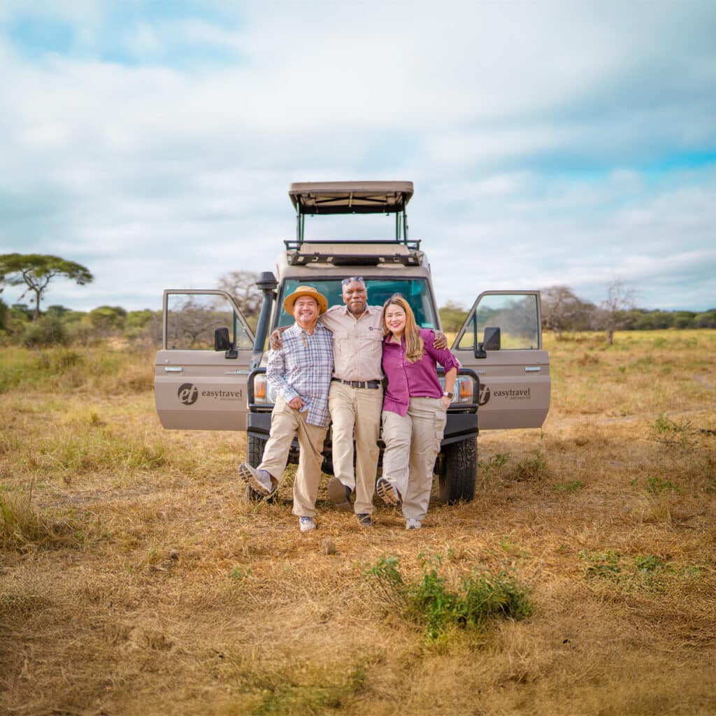 Three travelers pose in front of an easy travel safari vehicle in a grassy landscape, showcasing adventure and camaraderie.