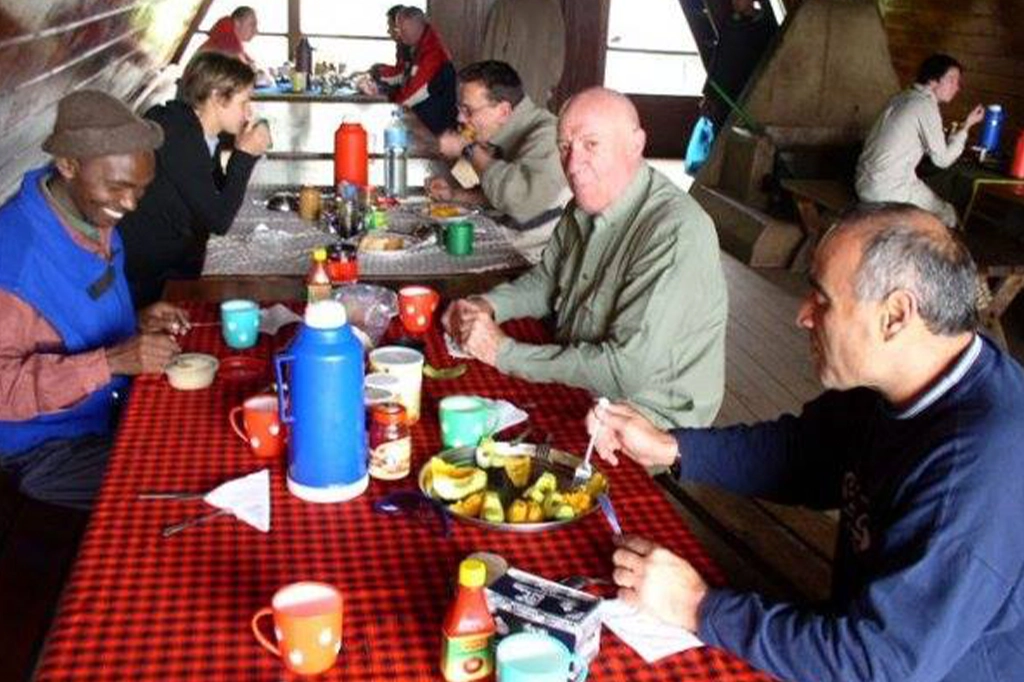 Un primo piano di un gruppo eterogeneo di escursionisti e membri della squadra di montagna seduti attorno a un tavolo a scacchi rossi e neri, che mangiano un pasto all'interno di una capanna