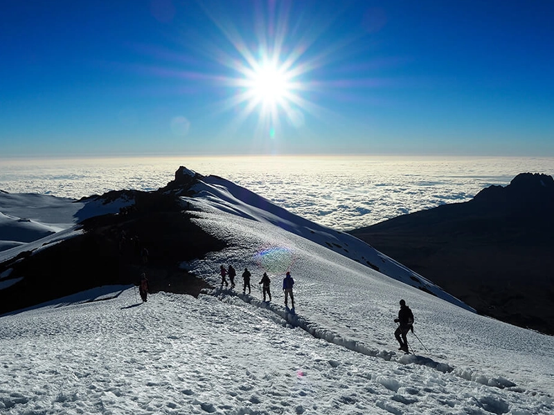 A line of hikers on the snowy slopes of Mount Kilimanjaro above the clouds, with a bright sunburst overhead.