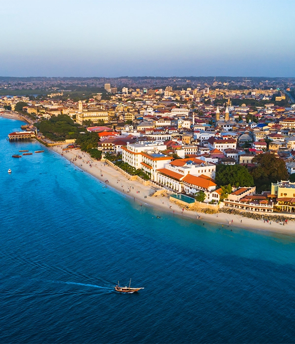 An aerial view of the historic Stone Town in Zanzibar, showing crowded buildings along a white-sand beach and a traditional dhow boat sailing on the turquoise ocean.