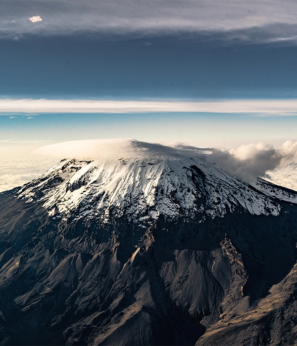 An aerial view of the snow capped Uhuru peak of Mount Kilimanjaro rising dramatically from dark volcanic slopes