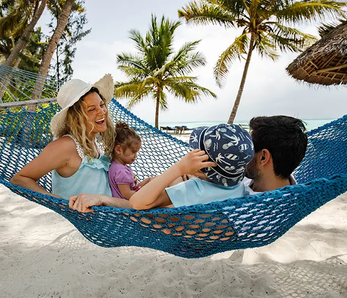 一家人穿着夏装，戴着帽子，在棕榈树环绕的热带海滩上，躺在吊床上享受阳光明媚的一天。.