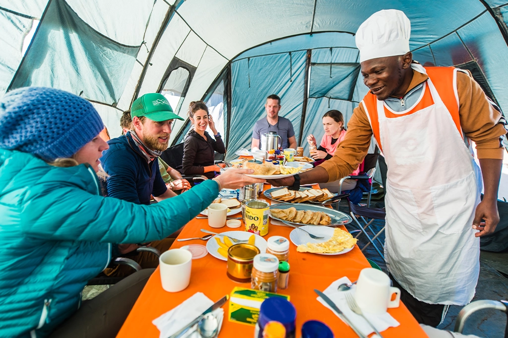 Easy Travel Mountain Chef serveert een maaltijd aan blije wandelaars in een eettent op de Kilimanjaro Easy Travel Mountain Chef serveert een maaltijd aan blije wandelaars in een eettent op de Kilimanjaro