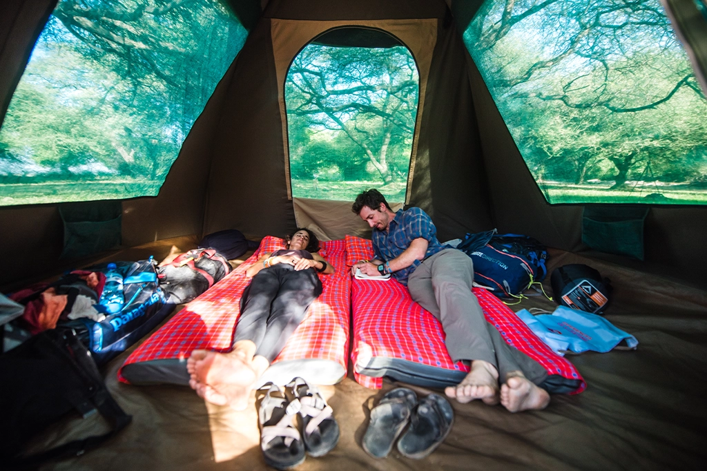 Dentro de una tienda de campaña en el campamento del lago Eyasi, una pareja yace sobre colchonetas para dormir, mientras el hombre escribe en un cuaderno y el bosque es visible a través de las ventanas de malla de la tienda.
