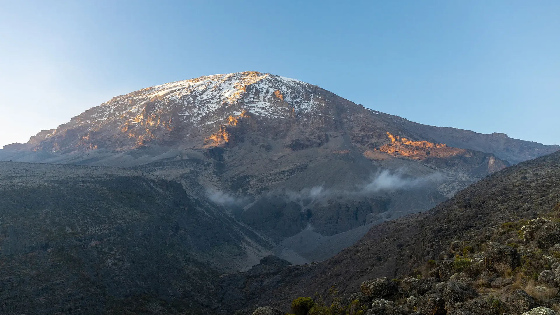 Snow-capped Mount Kilimanjaro rises above rocky mountain slopes at sunrise during Machame Kilimanjaro & Great Migration Safari.