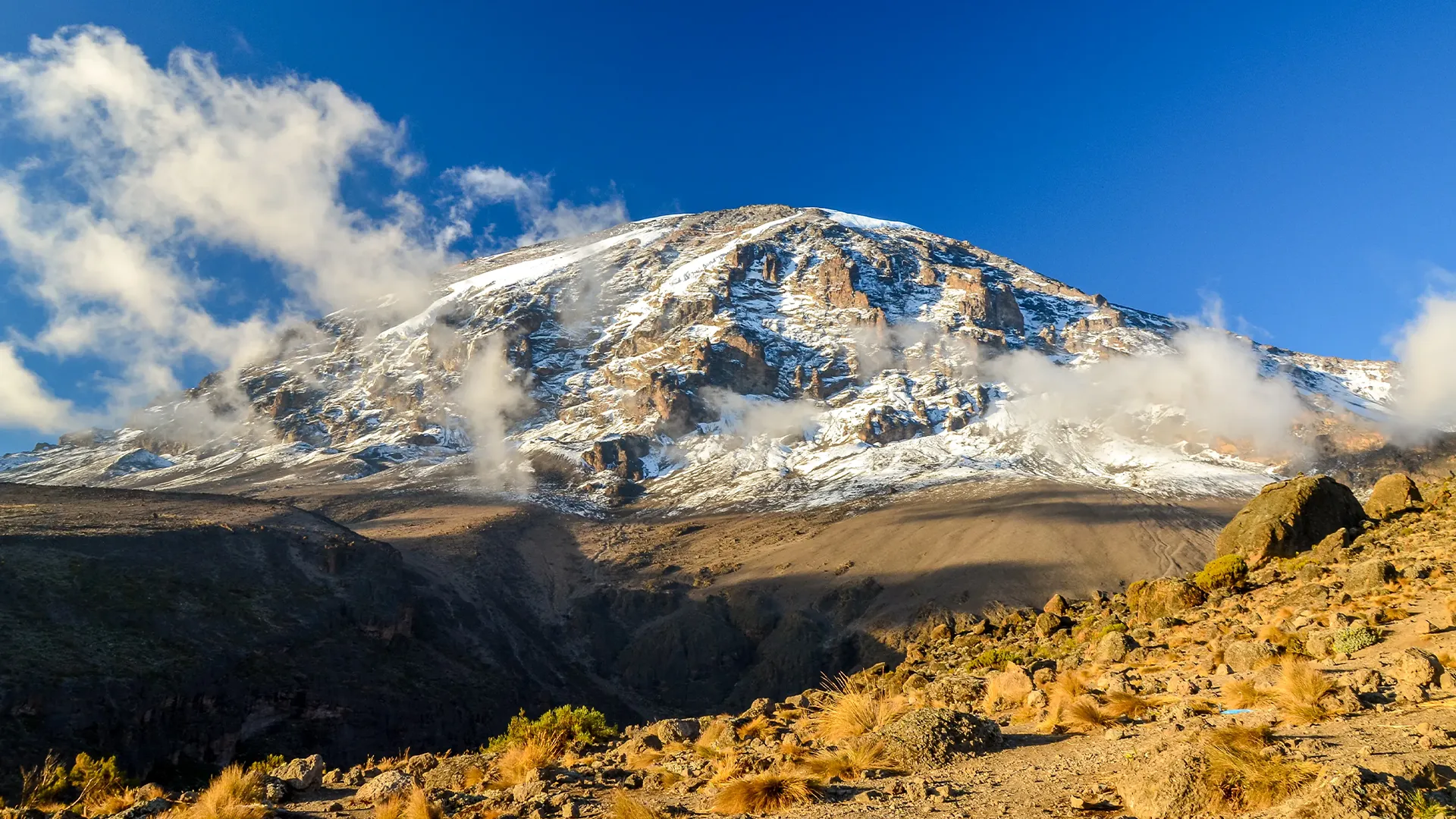 Snow-capped Mount Kilimanjaro rises above rocky slopes under a clear blue sky during Machame Kilimanjaro Great Migration Safari & Zanzibar Tour.