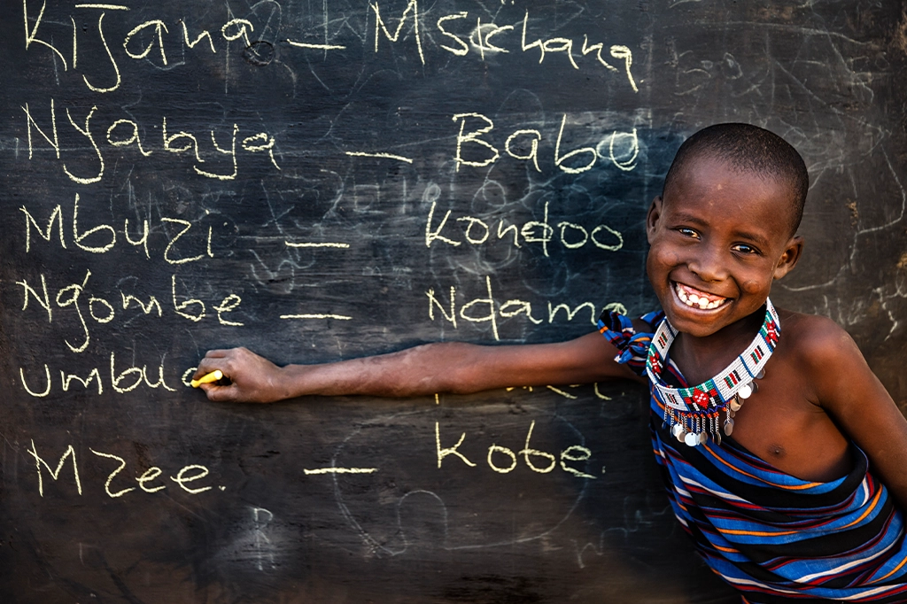 一位面带微笑的马赛族儿童,脖子上戴着串珠项链,正在黑板前用斯瓦希里语写字。 一位面带微笑的马赛族儿童,脖子上戴着串珠项链,正在黑板前用斯瓦希里语写字。