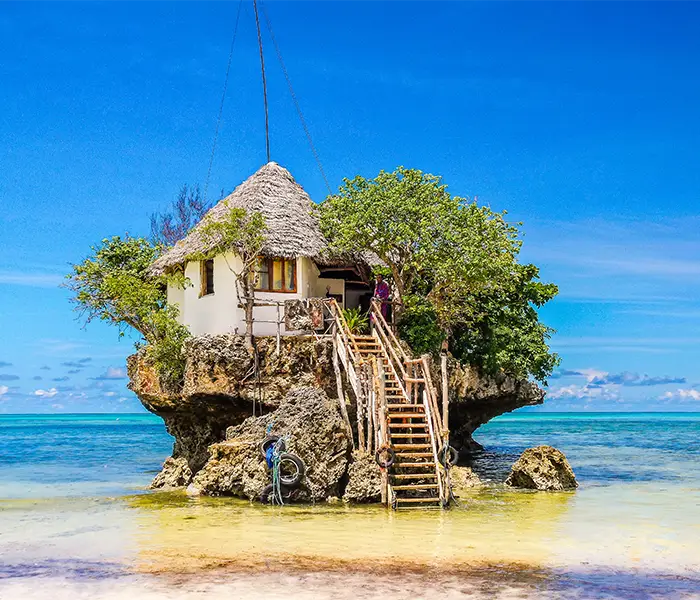 海边餐厅建在岩石上，屋顶是茅草顶，周围环绕着碧绿的海水和湛蓝的天空。.