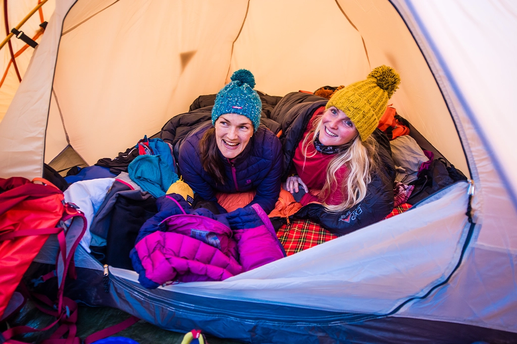 Deux femmes souriantes, coiffées de bonnets et vêtues de vestes d'hiver, sont allongées dans des sacs de couchage à l'intérieur d'une tente.